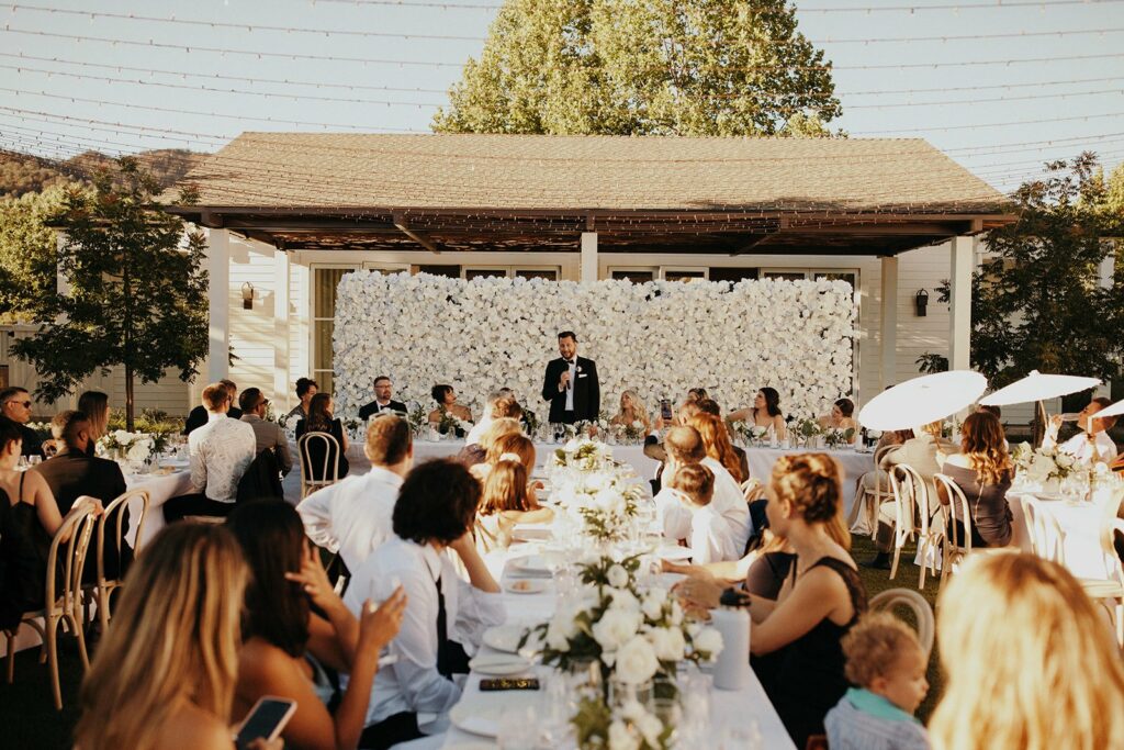 A wedding reception with guests seated at outdoor tables, a flower-adorned backdrop, and a man giving a speech in front of the wedding party on a raised platform.
