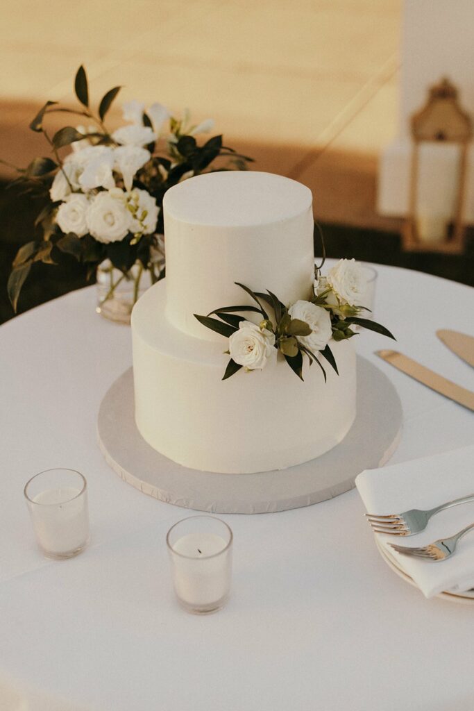 A two-tiered white cake adorned with white flowers and green leaves, placed on a round table with candles, a floral arrangement, napkins, and cutlery.