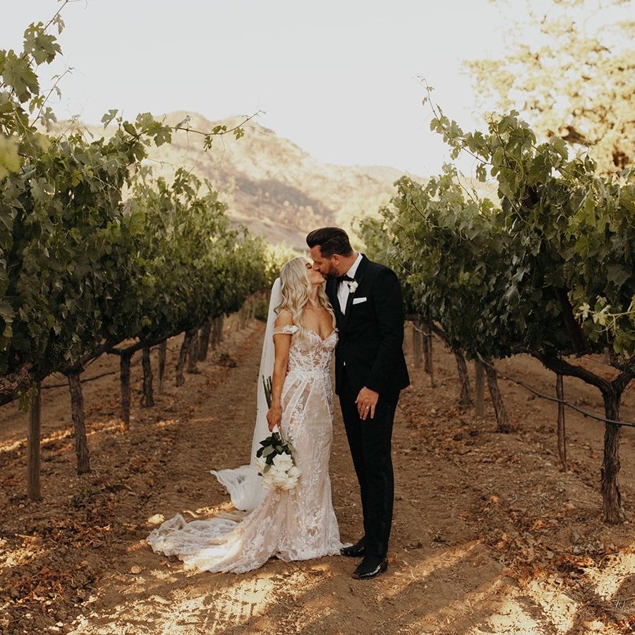 A bride and groom share a kiss while standing in a vineyard, surrounded by rows of grapevines. The bride holds a bouquet of white flowers.