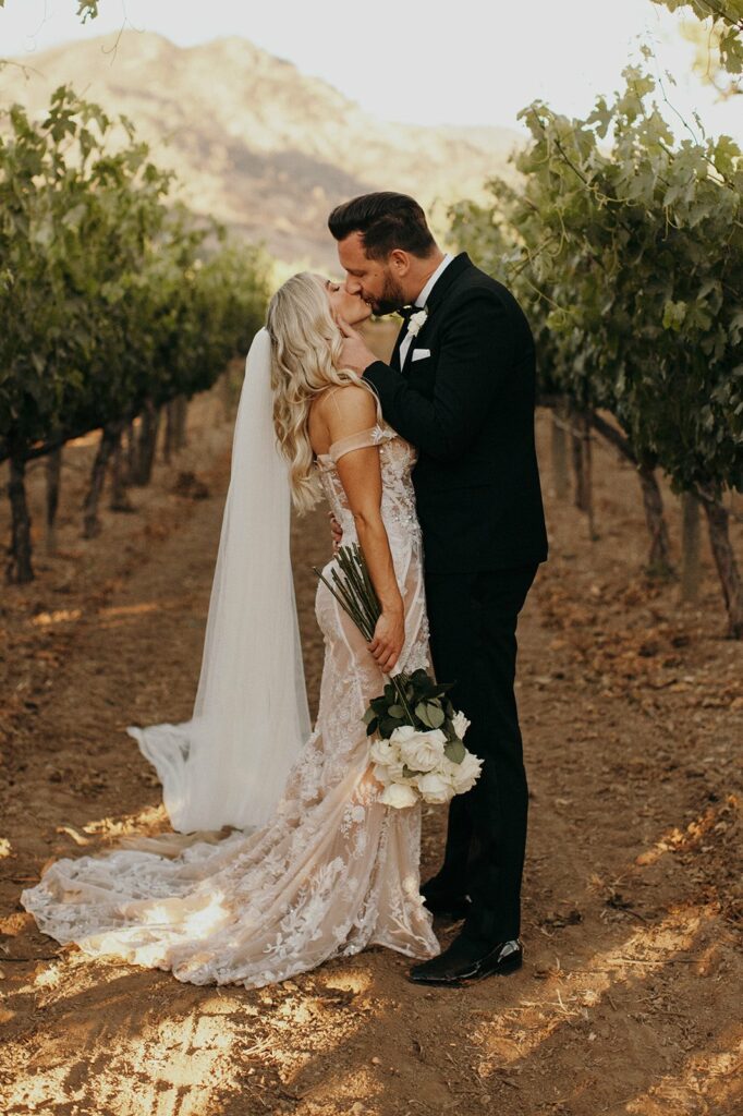 A bride and groom share a kiss in a vineyard. The bride is holding a bouquet of white flowers and wears an off-the-shoulder lace dress with a long train. The groom wears a black suit.