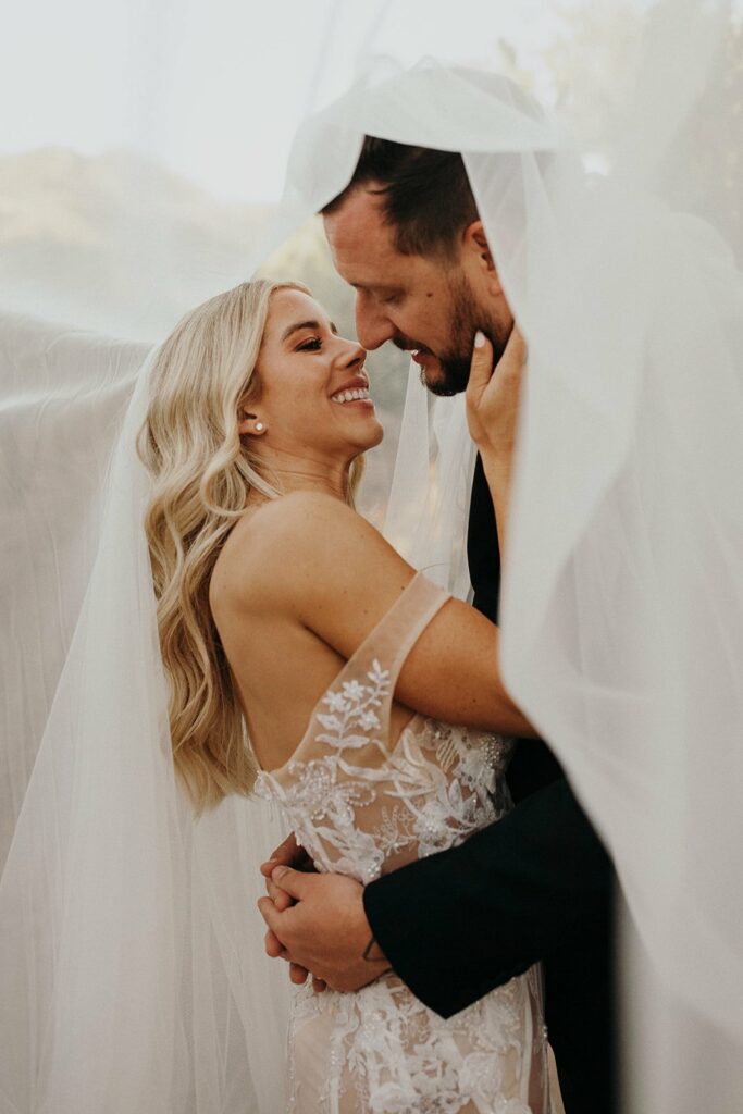 A bride and groom stand close under a veil, smiling at each other. The bride has long blonde hair and is wearing a white lace dress. The groom has short hair and is wearing a dark suit.