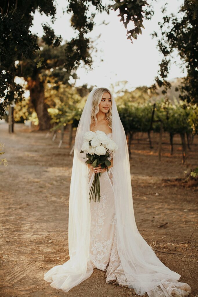 A bride in a white wedding dress and long veil stands outdoors holding a bouquet of white roses, with trees and a vineyard in the background.