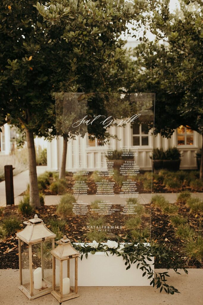 A transparent acrylic wedding seating chart is displayed outdoors among trees and greenery, listing the names and table assignments. Two lanterns with candles are placed at the base.