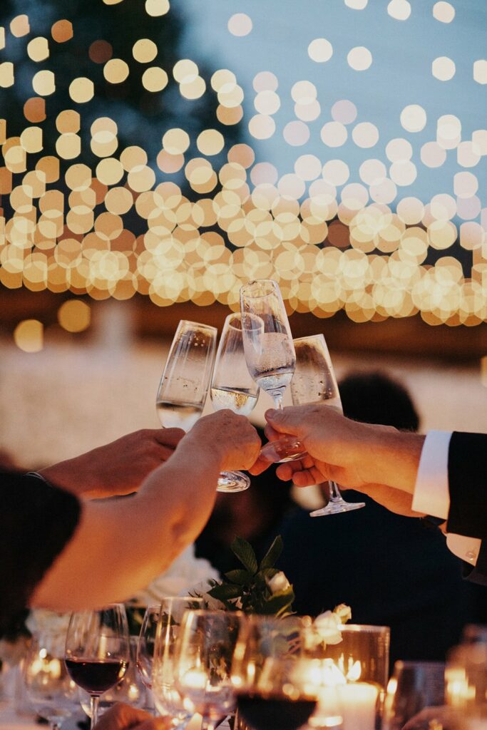 Several hands clinking champagne glasses in a toast with a backdrop of string lights, surrounded by candles and wine glasses on a table.