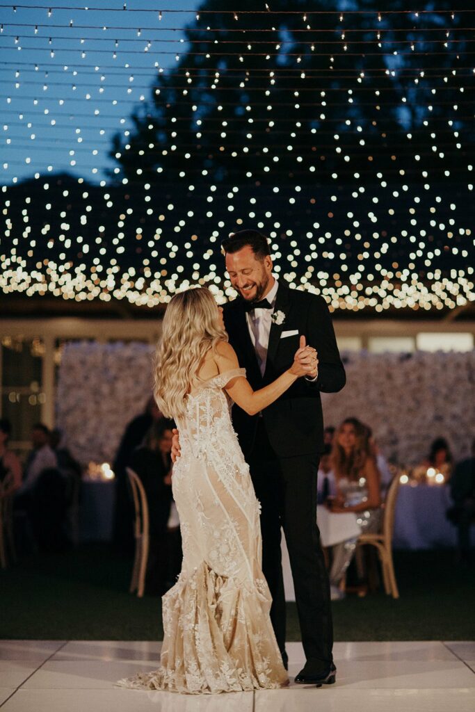 A couple dances under string lights at an outdoor event during the evening. The man is wearing a black suit and the woman is in a white dress. People are seated in the background.