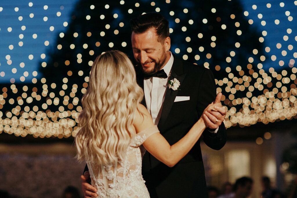 A couple dressed in formal wedding attire share a dance under a canopy of string lights.