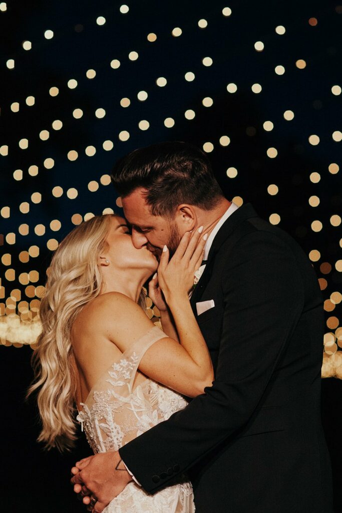 A couple embraces and kisses at night under a backdrop of numerous small, glowing lights. The woman wears a lace dress, and the man is in a dark suit.