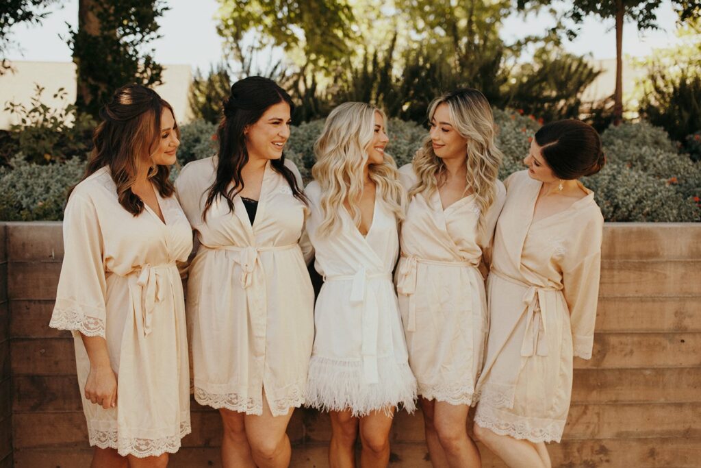 Five women in robes stand together outdoors by a concrete wall, with leafy green trees and shrubs in the background. Four of the women wear matching beige robes, and the woman in the center wears a white robe.