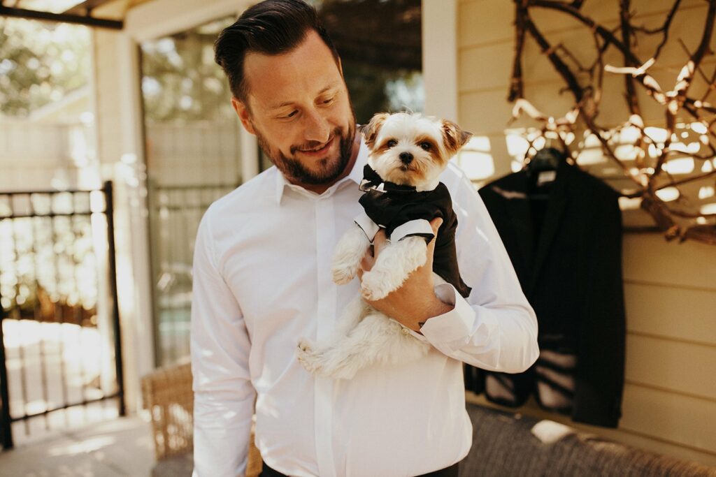 A man in a white shirt holds a small dog dressed in a tuxedo. They are standing outside near a wall with hanging branches.