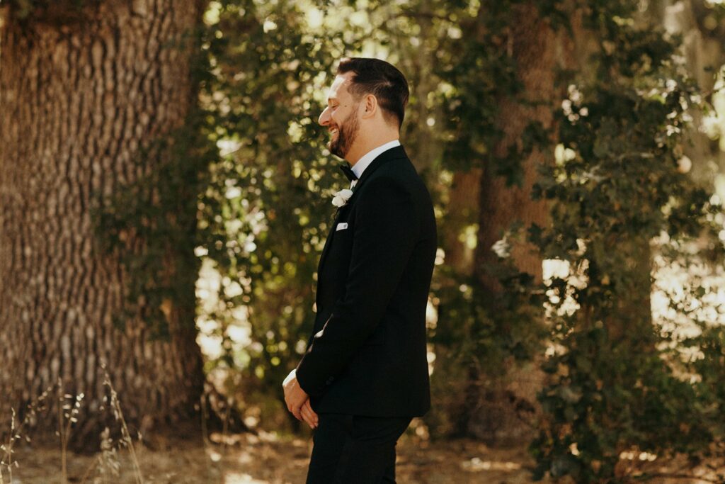 A man in a black suit with a white boutonnière stands outdoors in a wooded area, smiling.