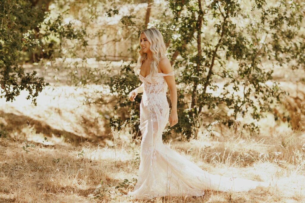 A woman in a lacy white dress walks through a sunlit forested area with dry grass and trees.