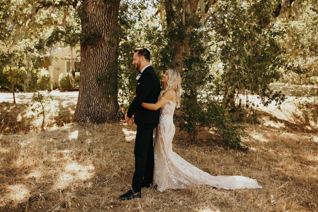 A woman in a wedding dress is standing behind a man in a suit outdoors, both under trees in a sunny, wooded area with dry grass.