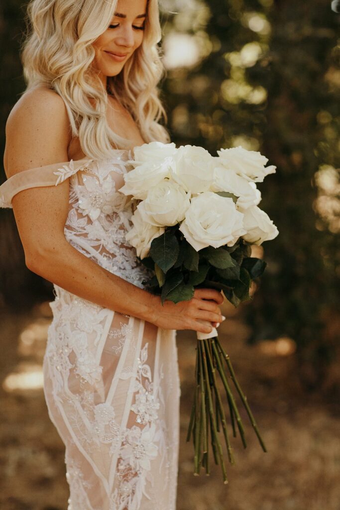 A woman in a lace wedding dress holds a bouquet of white roses, standing outdoors in a natural setting.