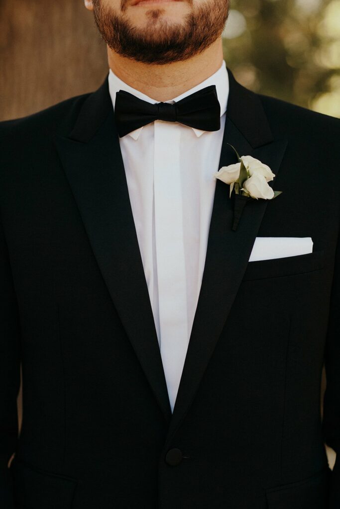 A man in a black tuxedo with a white shirt, black bow tie, and white rose boutonnière is shown from the neck down.