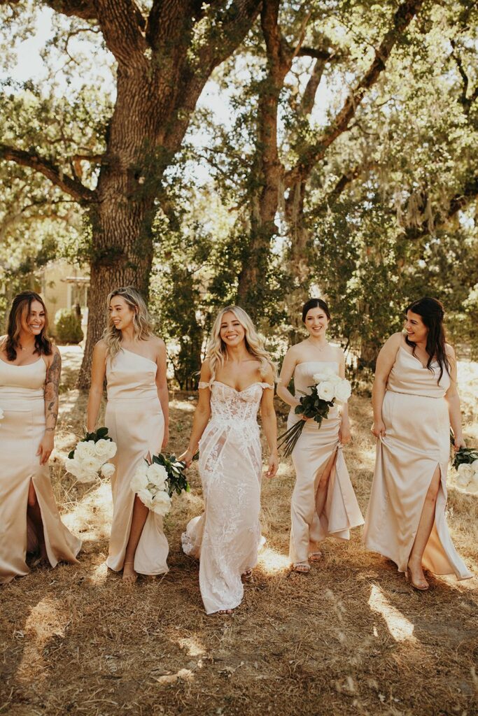A bride in a white lace dress walks outdoors with four bridesmaids in beige dresses, all holding white flower bouquets. Trees and greenery are in the background.