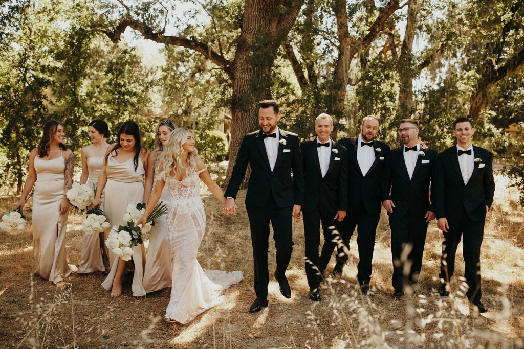 A bride and groom walk hand-in-hand outdoors, surrounded by bridesmaids in cream dresses and groomsmen in black tuxedos, all smiling and conversing.