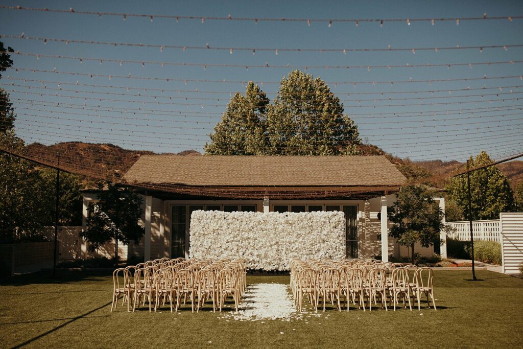 An outdoor wedding ceremony setup with rows of wooden chairs facing a white floral backdrop. Overhead string lights are hung above the area. Trees and a building are in the background.