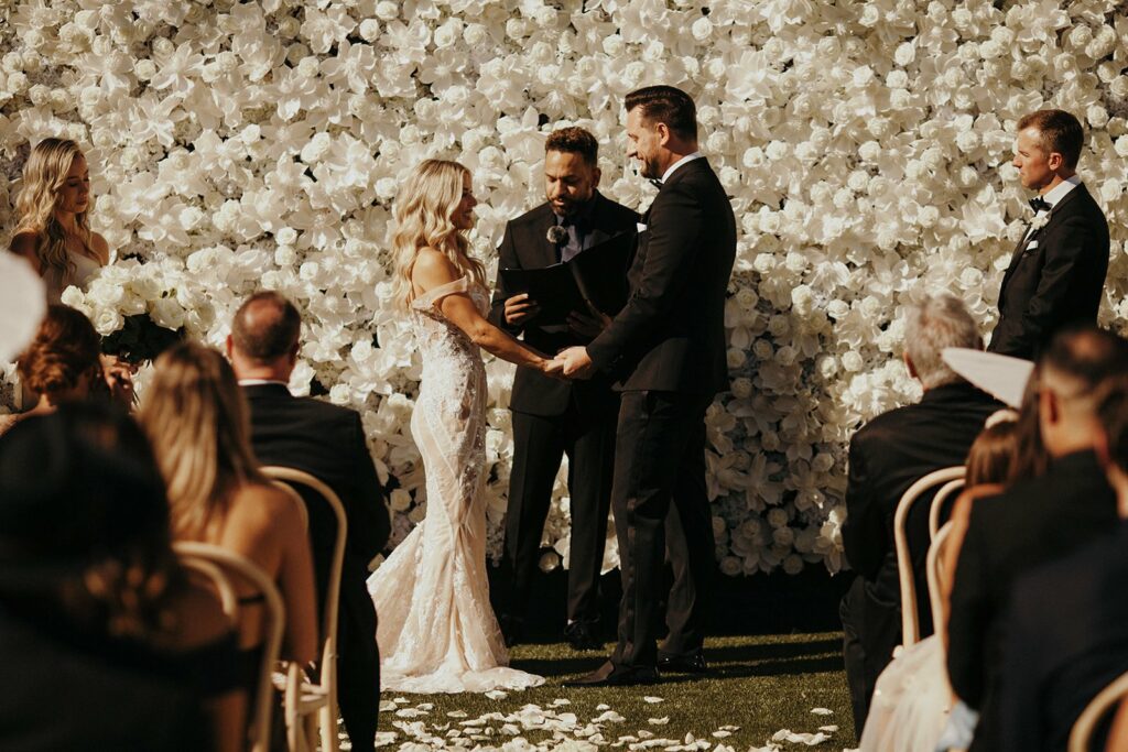 A couple exchanges vows during an outdoor wedding ceremony in front of a wall adorned with white flowers. Guests watch seated on both sides of the aisle.