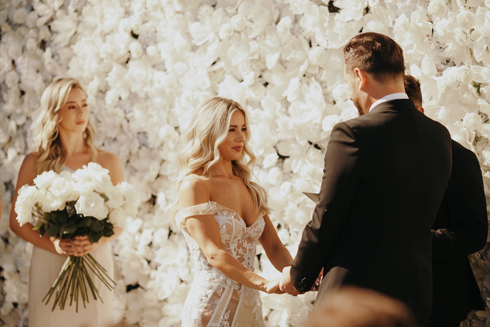 A couple stands facing each other and holding hands during a wedding ceremony, with a backdrop of white flowers. A bridesmaid holding a bouquet of white flowers stands in the background.