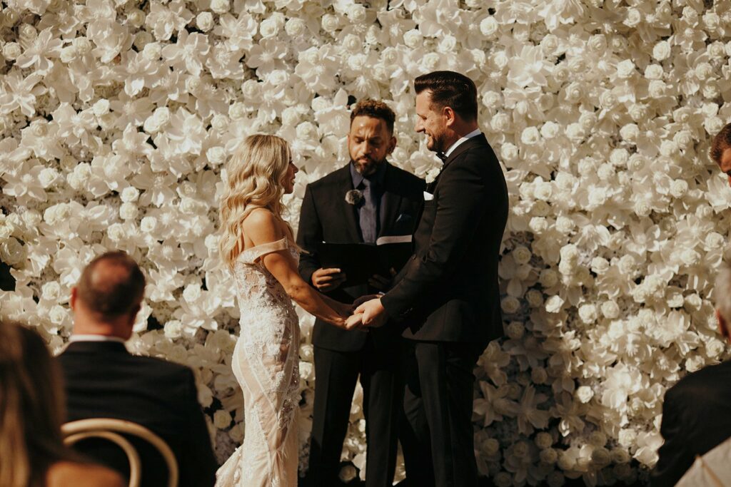 A couple stands holding hands during their wedding ceremony in front of a white flower wall, while an officiant reads. Guests are seated and watching the ceremony.