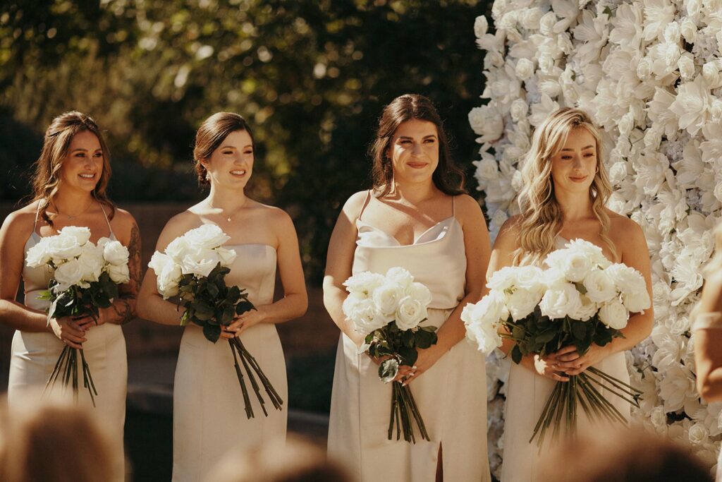Four bridesmaids in light-colored dresses stand holding white bouquets in front of a floral backdrop during an outdoor event.