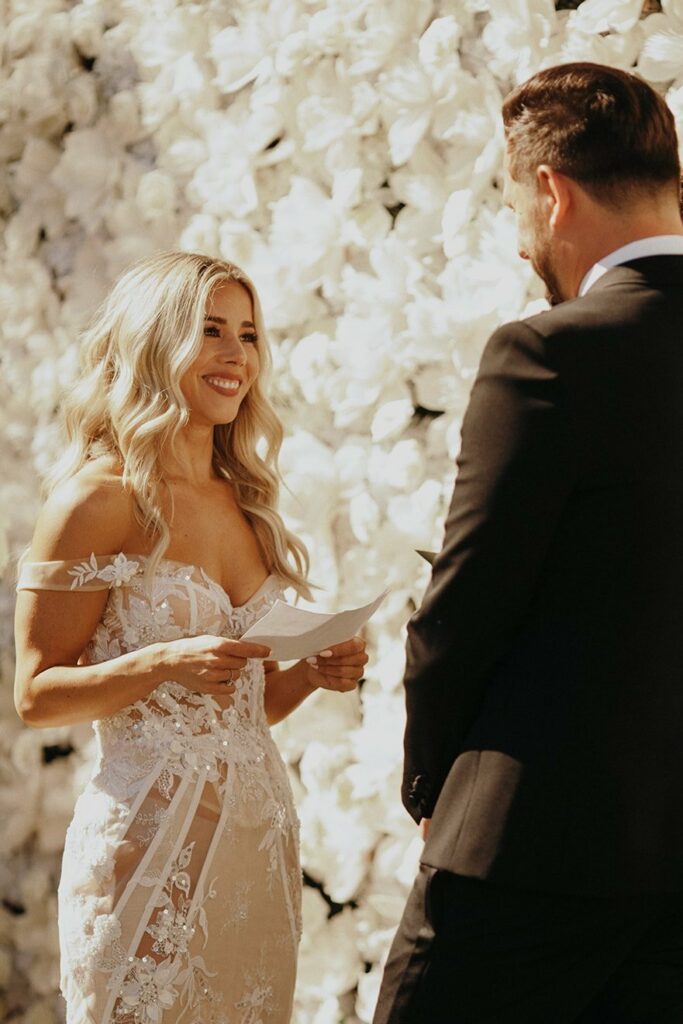 A bride in a white lace dress reads vows to a groom in a black suit in front of a wall of white flowers.