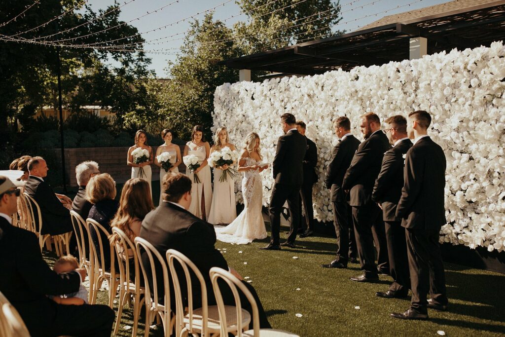 A bride and groom stand at the altar during an outdoor wedding ceremony, with bridesmaids and groomsmen in formal attire. Guests are seated on chairs, and white floral decorations adorn the background.
