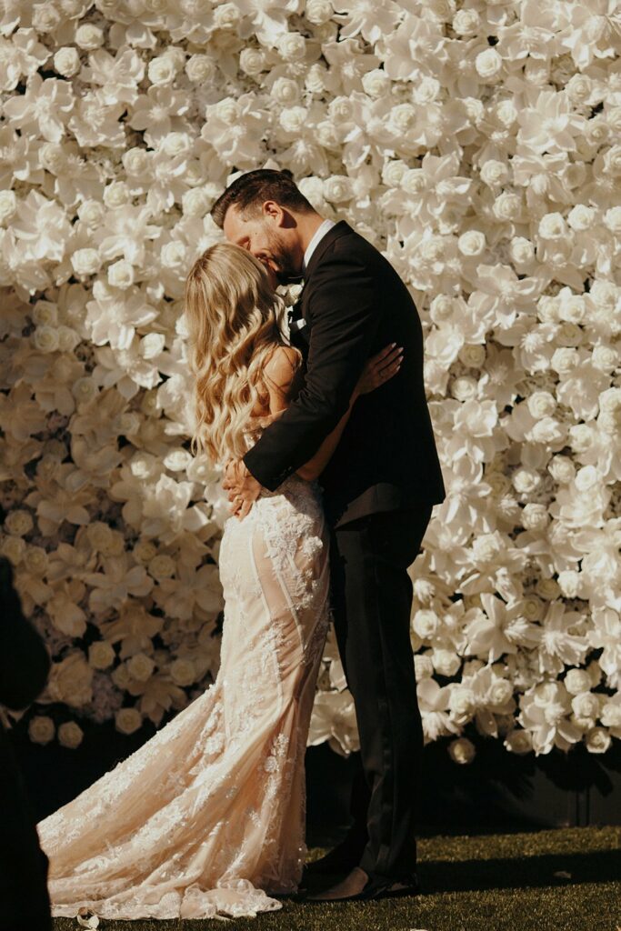 A bride and groom in elegant attire share a kiss in front of a wall decorated with numerous white flowers in an outdoor setting.