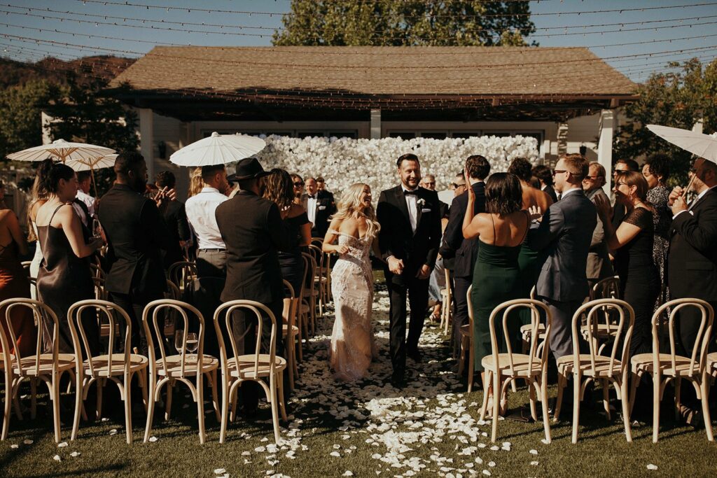 A newly married couple walks down the aisle outdoors, surrounded by seated and standing guests, some holding white umbrellas. Flower petals are scattered on the ground.
