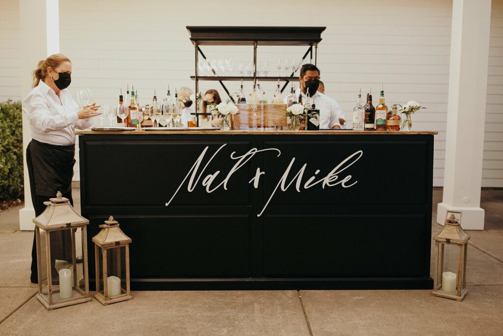 Two bartenders stand behind a black bar with "Nat & Mike" written on it, preparing drinks. The bar is decorated with flowers and surrounded by lanterns with candles.