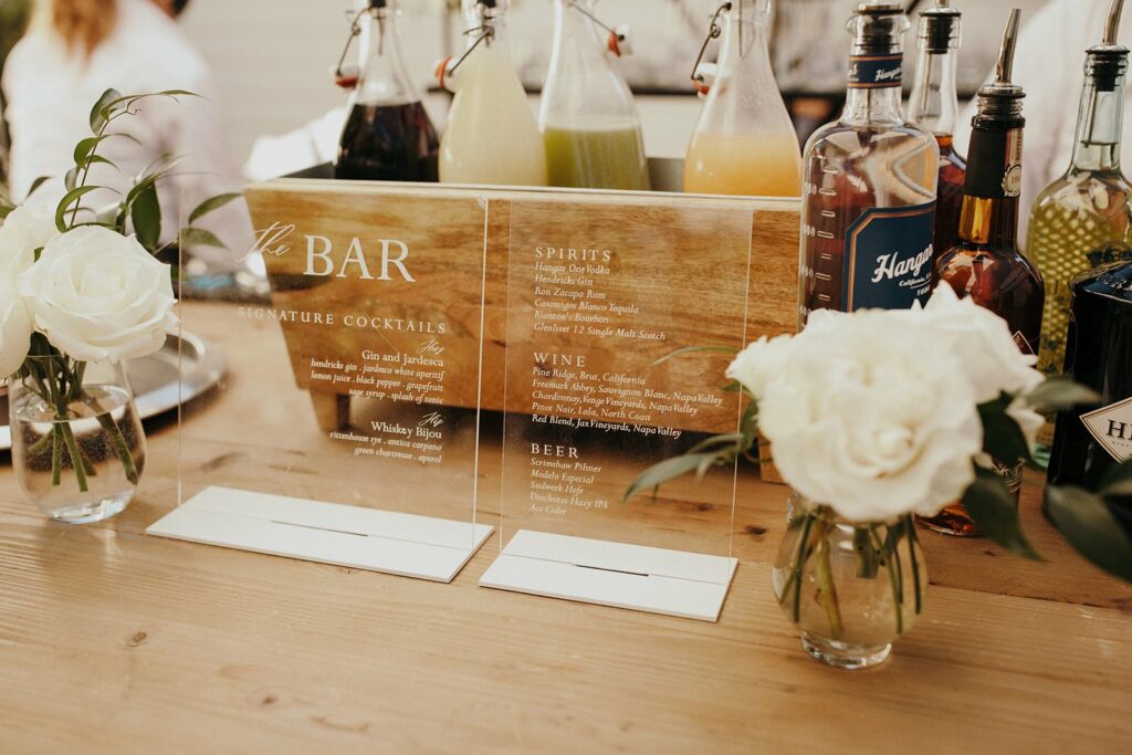 A wooden bar menu with a list of cocktails, spirits, wine, and beer. Various bottles and mixers, as well as white roses in vases, are displayed on a wooden table.
