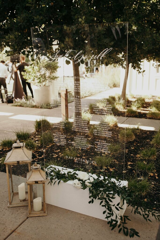 A clear acrylic wedding seating chart with guest names is displayed outdoors, leaning against greenery. Two wooden lanterns with candles and green foliage are placed at the base.
