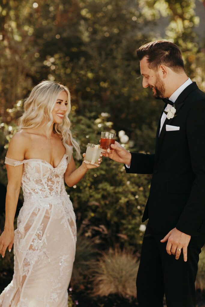 A bride in a lace gown and a groom in a black tuxedo toast with drinks in an outdoor setting.