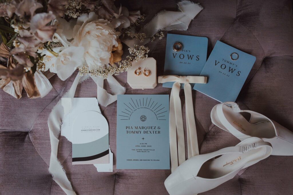 A wedding invitation and vow booklets are displayed on a purple surface, accompanied by white flowers, a pair of white shoes, two gold rings in a box, and a ribbon.