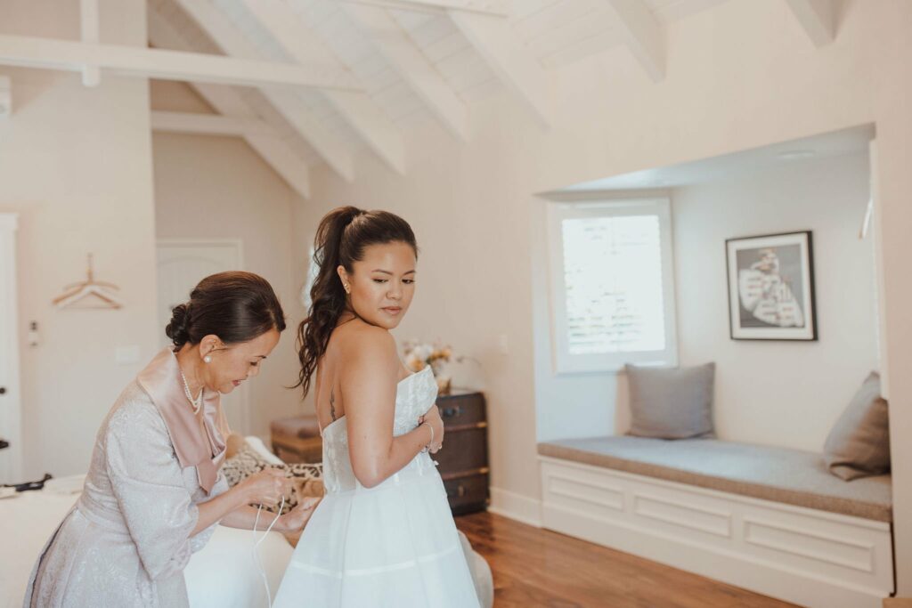 A bride, wearing a white strapless dress, is receiving assistance from another woman in a light-colored dress, inside a well-lit room with vaulted ceilings.