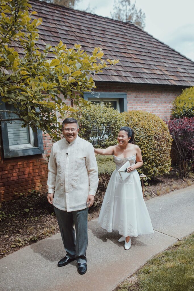 A bride and groom stand outside on a pathway. The bride is in a white dress holding an envelope, while the groom is in a cream-colored barong and gray pants, smiling. Brick building and bushes in the background.