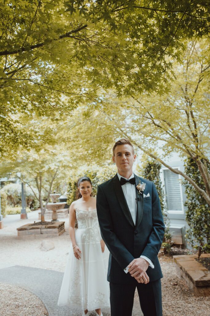 A bride in a white dress stands behind a groom in a dark suit and bow tie under a leafy canopy in an outdoor setting.