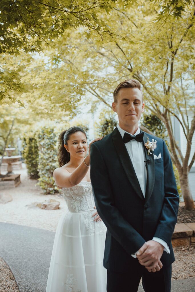 A bride in a white dress and a groom in a black suit stand outdoors. She gently touches his shoulder from behind, as they both prepare for a first look moment. Trees surround them.