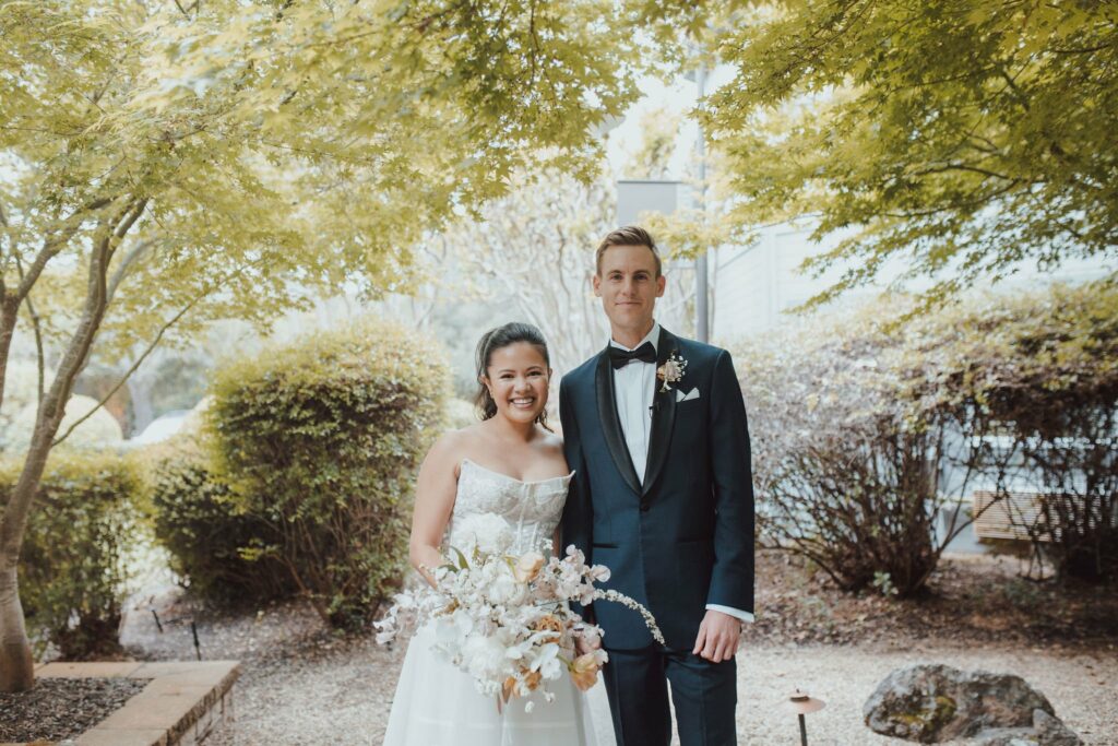 A bride in a white dress holding a bouquet stands next to a groom in a black tuxedo, both smiling, amidst greenery.
