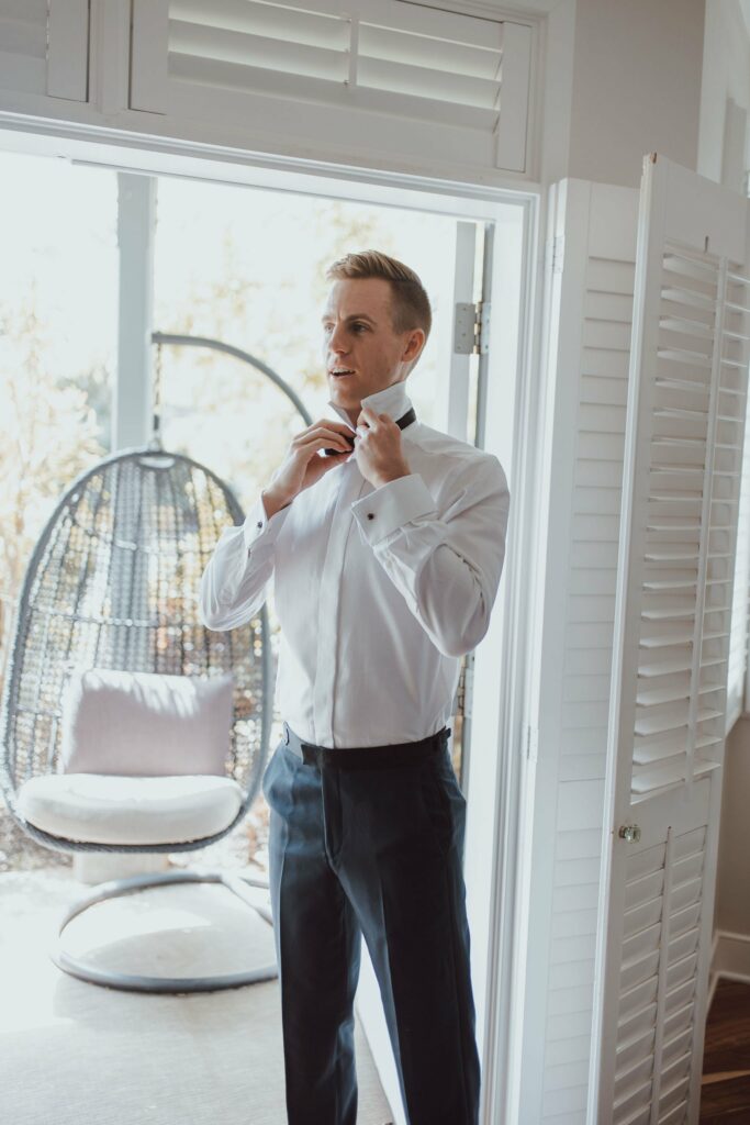 A man stands by a window adjusting his bow tie. He is dressed in a white shirt and black pants. A hanging chair and window with shutters are visible in the background.