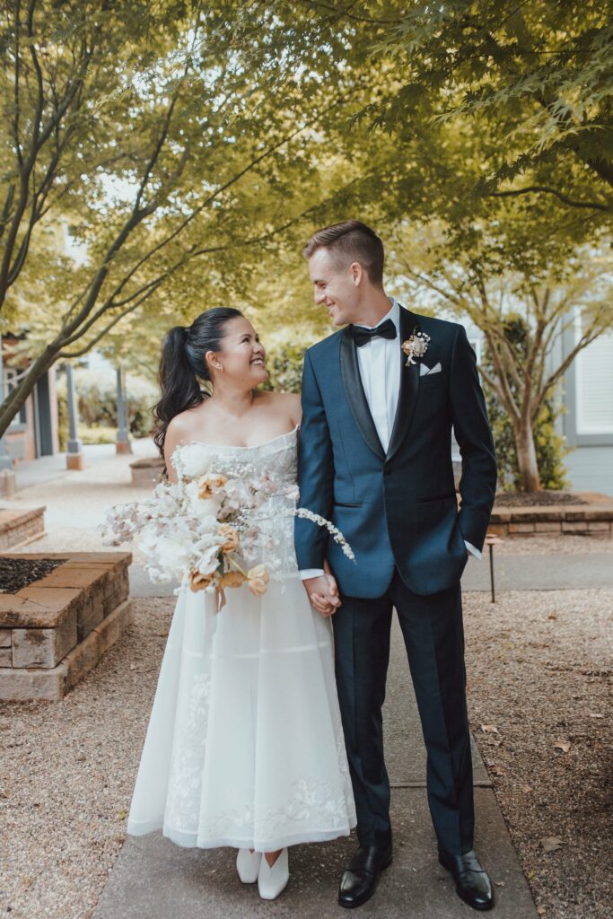 A couple dressed in wedding attire holds hands, smiling at each other outdoors under trees. The bride holds a bouquet, and both are walking on a gravel path.