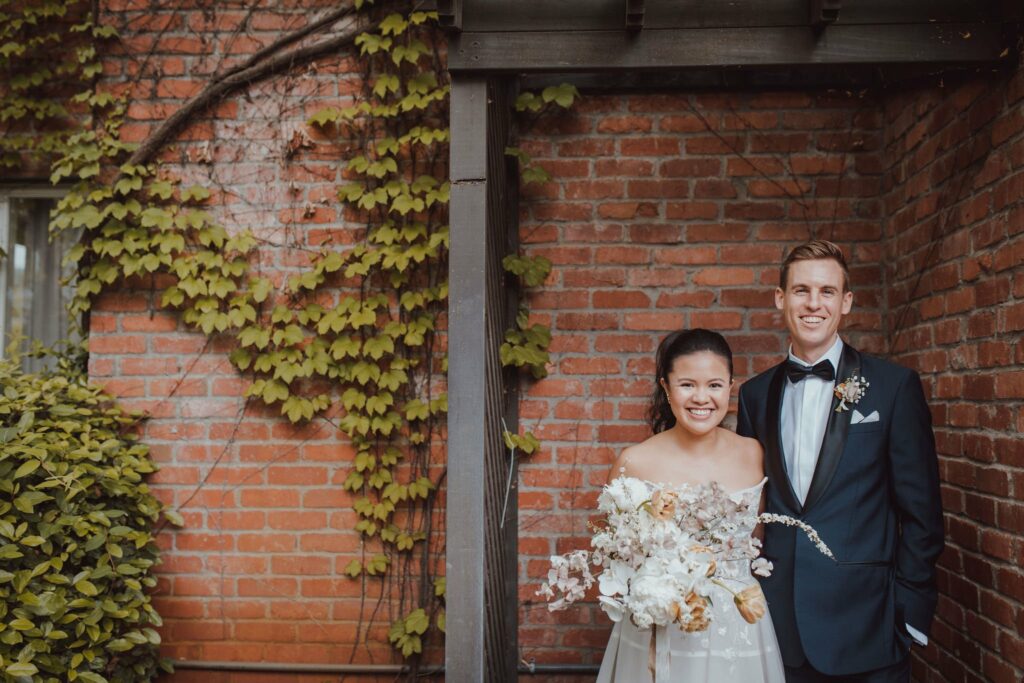 A couple in formal attire stands smiling under an outdoor wooden structure with brick walls and greenery in the background. The woman holds a bouquet of flowers.