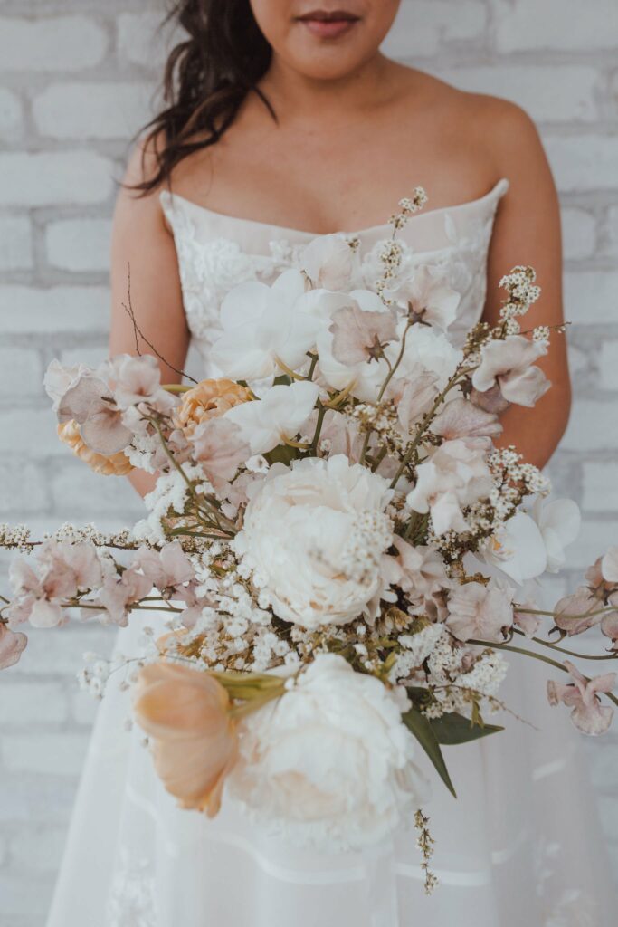 Close-up of a bride holding a bouquet of white and light brown flowers while wearing a strapless wedding dress. The background features a light gray brick wall.