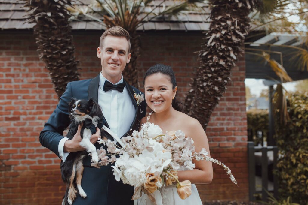 A bride and groom smile while holding a dog and a bouquet of flowers, standing in front of a brick wall and palm fronds.