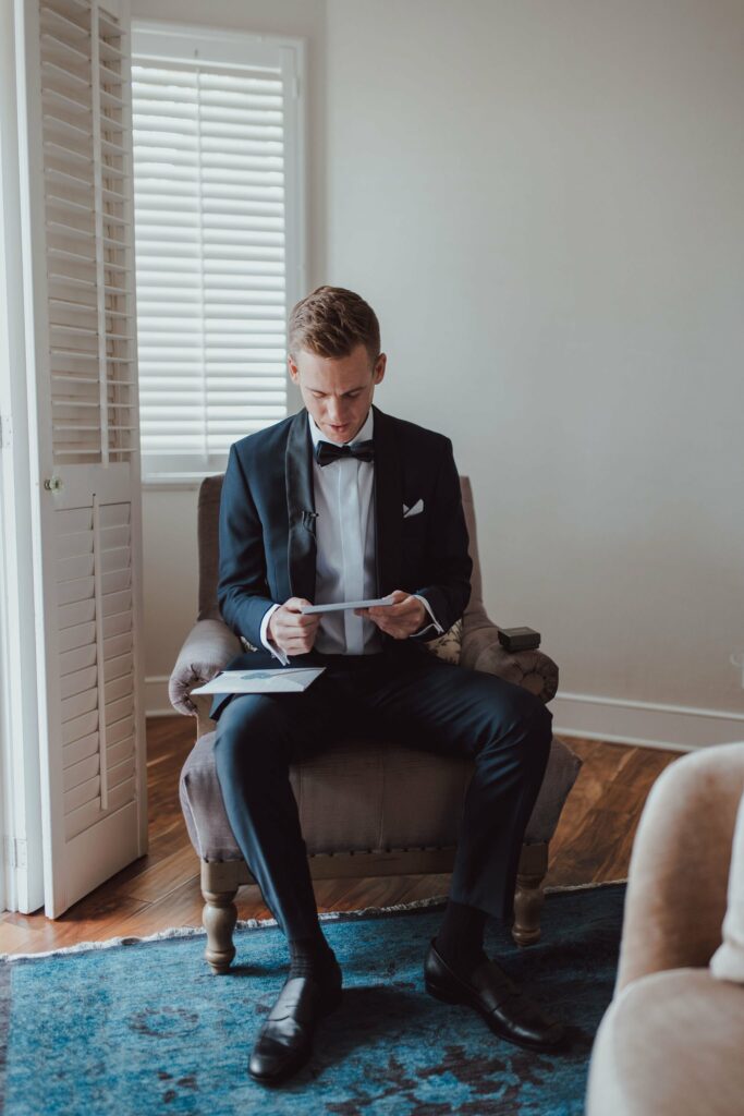 A man in a black tuxedo is seated on an armchair while looking at a piece of paper. He is in a room with closed white shutters and wooden flooring.