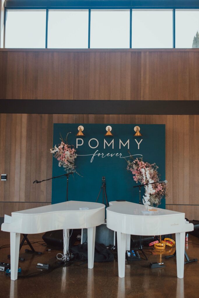 Two white grand pianos are set up in front of a backdrop reading "POMMY forever," adorned with floral arrangements and three hanging lights.