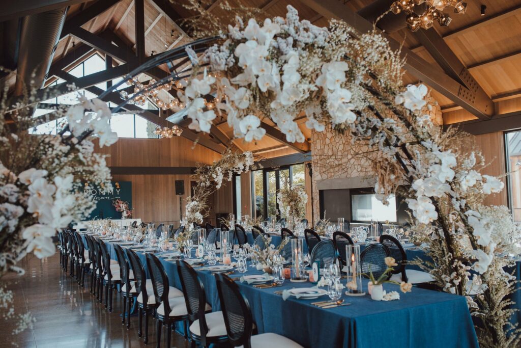 A decorated indoor dining area with long tables covered in blue tablecloths, white chairs, and floral arches adorned with white flowers. Large windows and wooden beams are visible in the background.