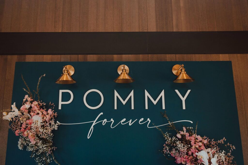 A sign that reads "POMMY forever" in white text on a blue background, decorated with pink and white floral arrangements and illuminated by three brass wall sconces.