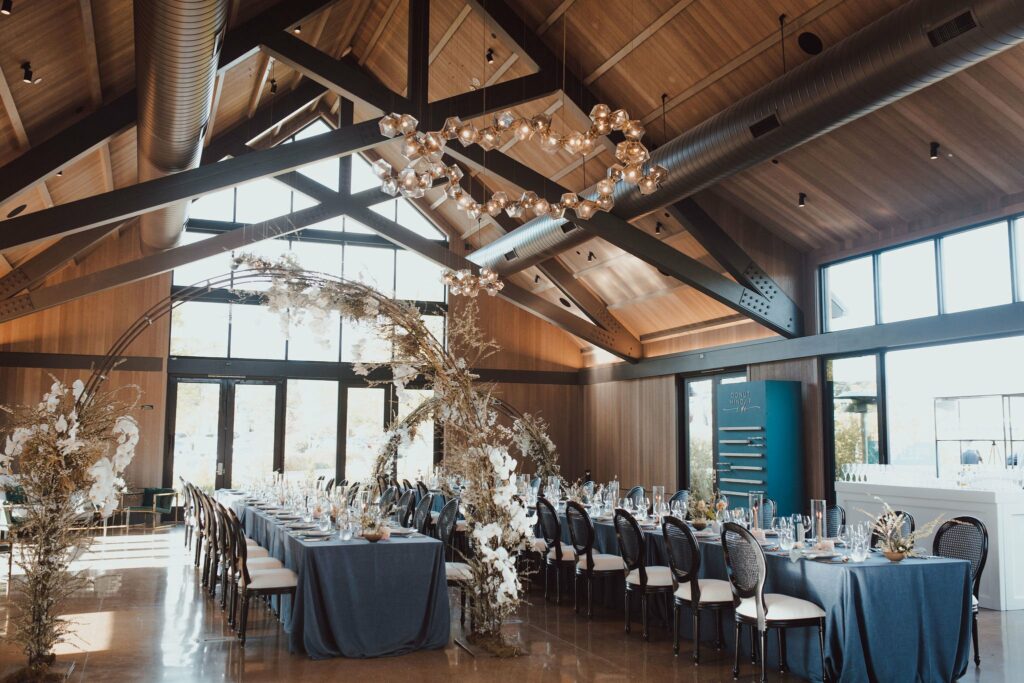A spacious event hall with high wooden ceilings features a long table set with blue tablecloths, chairs, and floral arrangements. Large windows on the side allow natural light to flood the room.