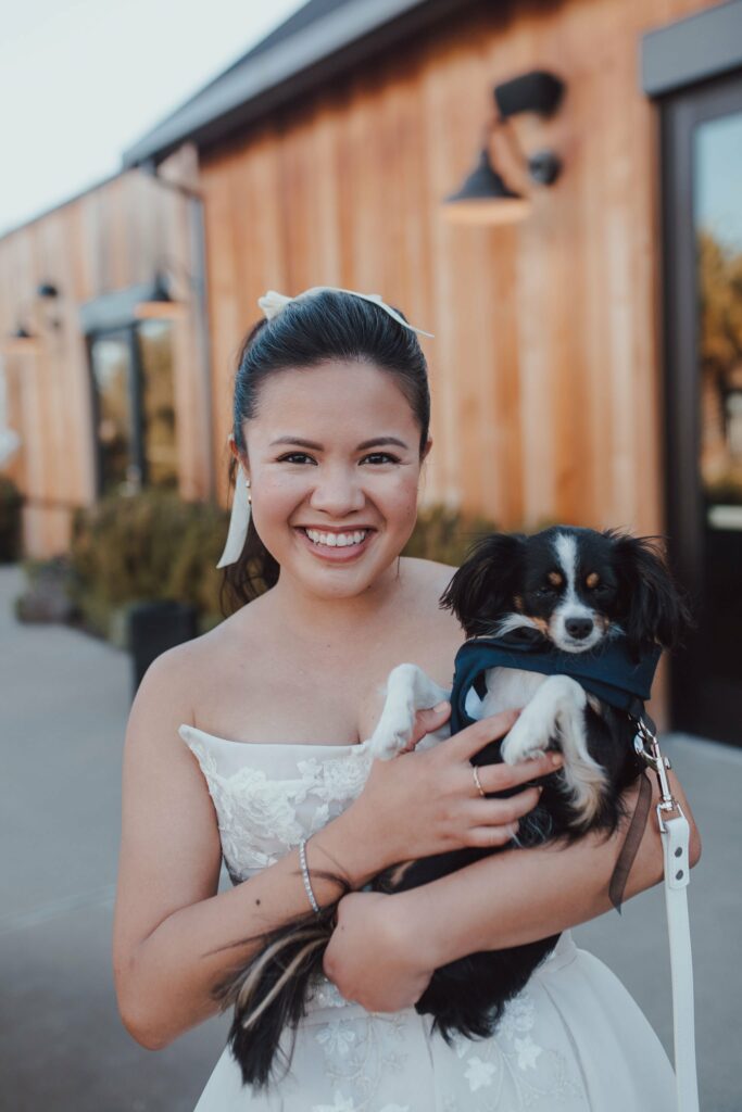 A woman in a white dress smiles while holding a small black and white dog in front of a wooden building.